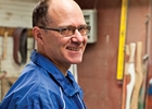 A smiling balding man stands with a drill ready to work on a canoe