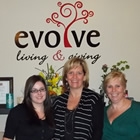 Leanne stands between her two employees behind the counter in front of the Evolve Living and Giving logo.