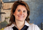 A smiling woman with mid-length brown hair stands against a painted landscape mural