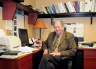 A balding man in a suit and tie with a huge smile sits at a desk