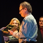 Teed, in blue shirt, receives plaque from announcer behind podium who is wearing a black dress as two people look on.