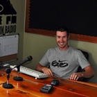 Scott sits behind a wide wood desk and station equipment.