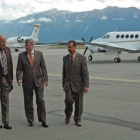 Three men in suits walk across the tarmac with two planes and mountains in the background.