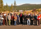 A group of 21 men and women stand on a site at the edge of town with forested mountains behind.