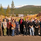 A group of 21 men and women stand on a site at the edge of town with forested mountains behind.