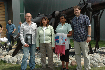 Two men and two women stand outdoors in front of a metal sculpture of a canoe.