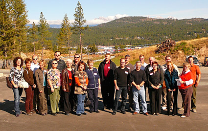 A group of 21 men and women stand on a site at the edge of town with forested mountains behind.